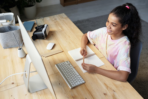Na imagem, uma menina está em sua mesa, usando o computador em sua rotina de estudos.