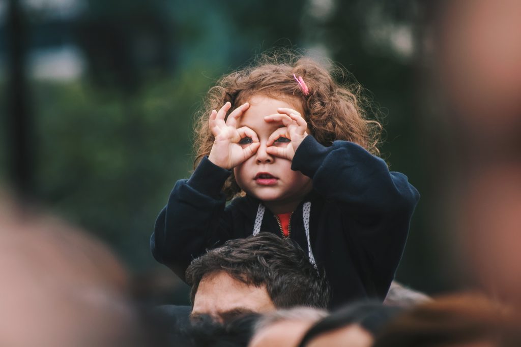 Criança menina sentada nos ombros dos pais com as mãos nos olhos, representando a psicomotricidade infantil.