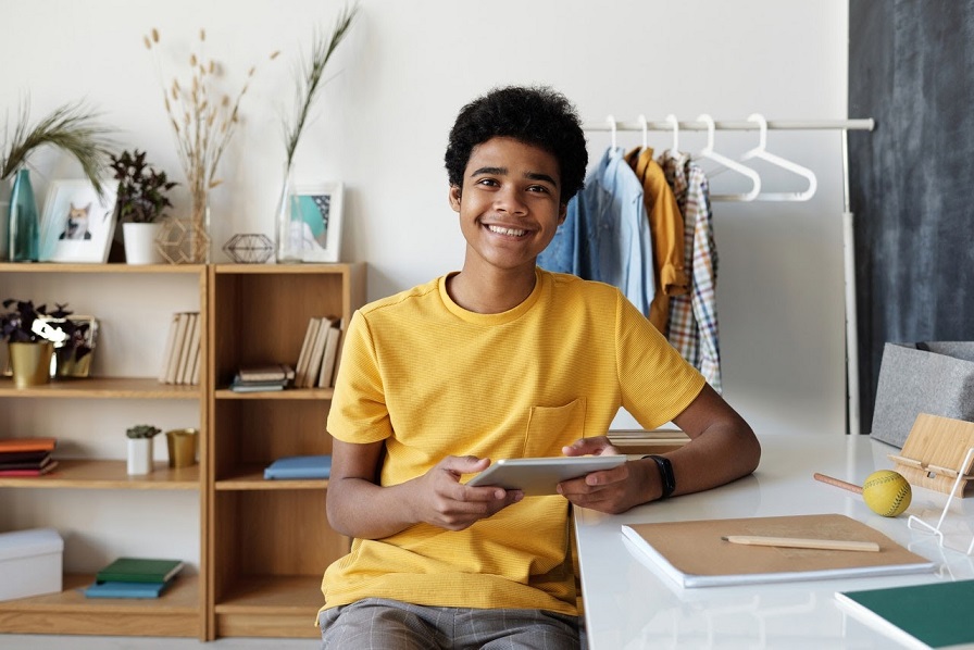 estudante com um tablet na mão, sentado com uma camiseta amarela, sorrindo para a câmera.