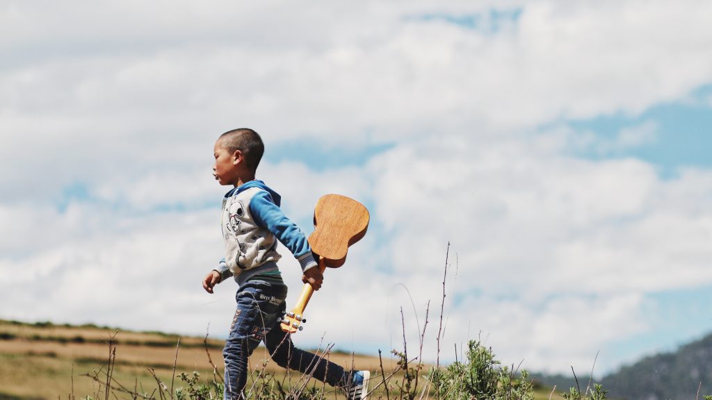 Menino criança andando por um campo com um violão em mãos.