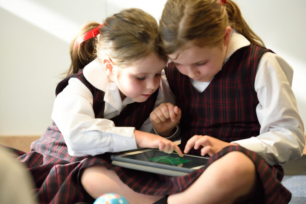 Duas meninas crianças com uniformes escolares brincando com um tablet demonstrando a importância do inglês.